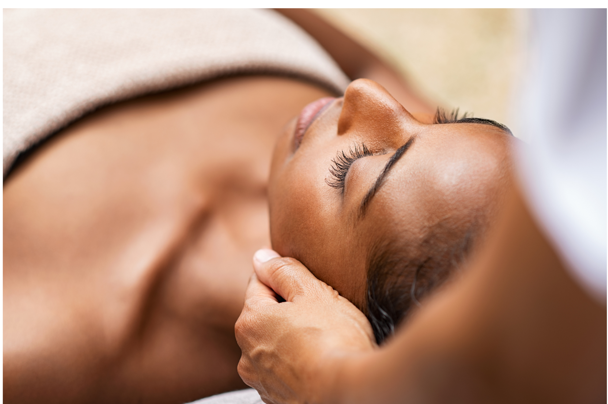 A woman receiving a facial treatment, lying down with her eyes closed, with another person's hand gently supporting her chin.