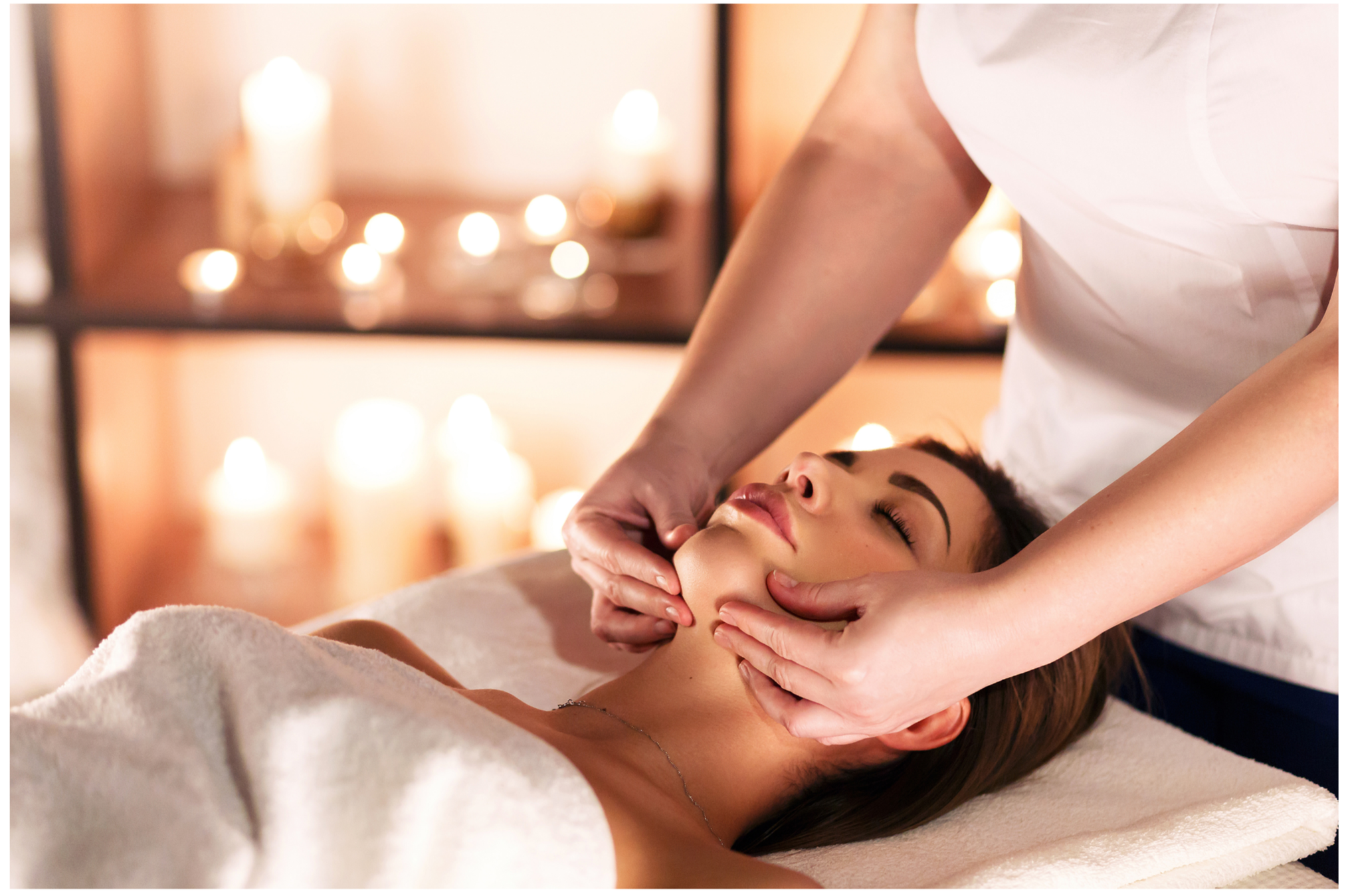 A woman is lying on a massage table during a facial massage, with eyes closed and a relaxed expression. A massage therapist is gently massaging her face with both hands. The background features soft, warm lighting and blurred candles, creating a calming spa atmosphere.