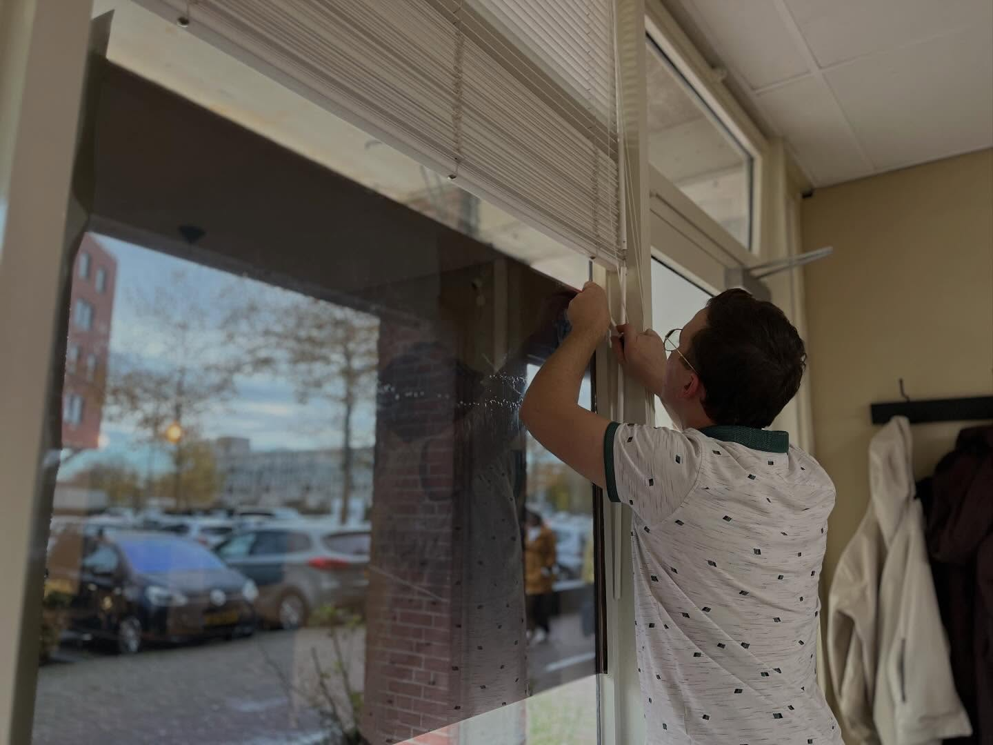A man wearing glasses and a face mask is placing a window film inside a room, with a view of parked cars and trees outside through the window.
