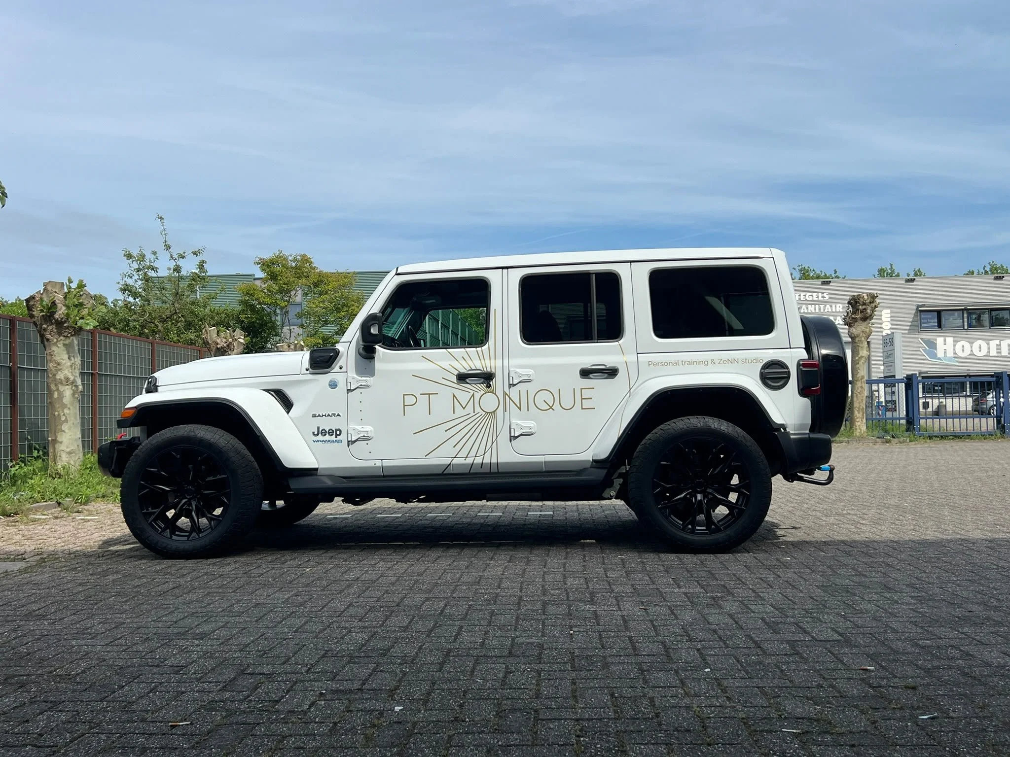 Wit Jeep Renegade met logo en tekst voor PT Monique op de zijkant, geparkeerd op een betegeld wegdek met bomen en een gebouw op de achtergrond.