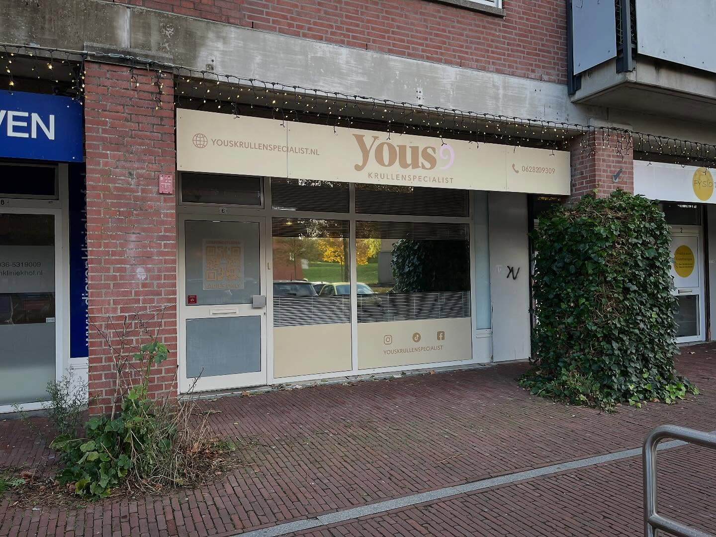 storefront with a sign that says 'Yous Krullenspecialist' with social media icons and contact information.