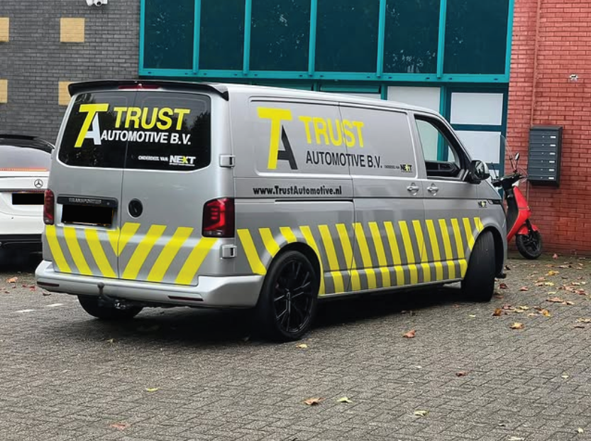 Silver van with yellow and black safety markings and large yellow text 'Trust' on the side, parked on a paved lot near a brick building, with a red scooter and other vehicles nearby.