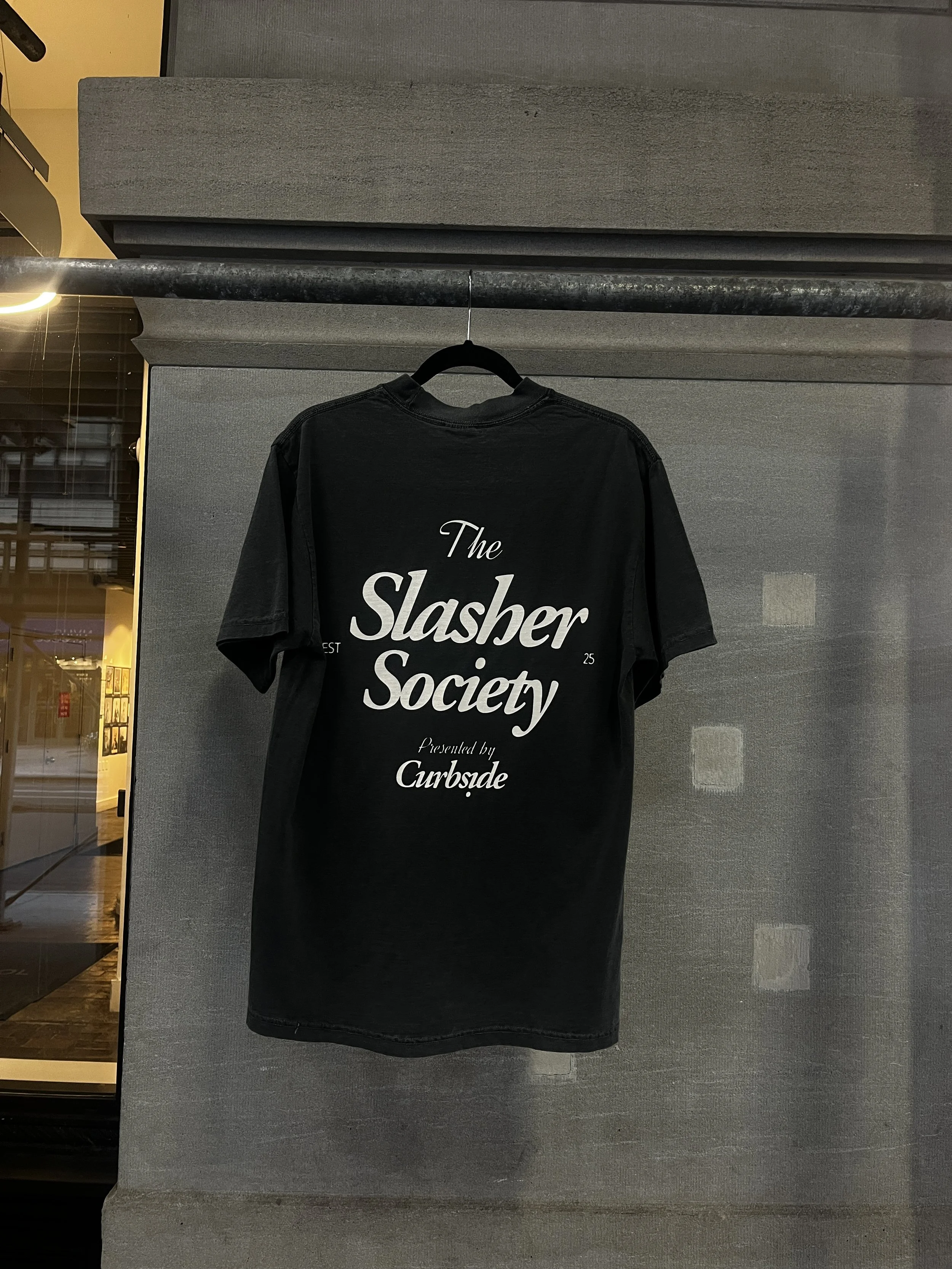 Black T-shirt with white text that reads 'The Slasher Society Presented by Curbside' hanging on a black hanger against a gray wall