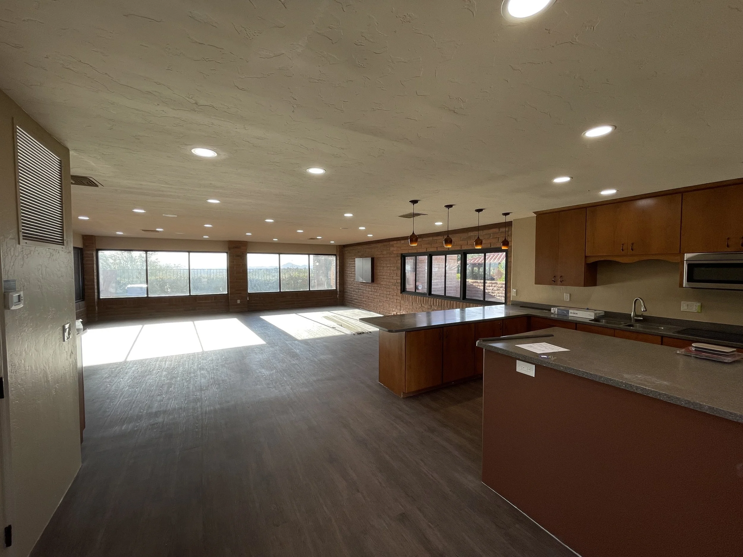 Interior of a spacious, open-plan living space with hardwood floors, large windows allowing natural light, a brick accent wall, and a kitchen area with wooden cabinets, a granite countertop island, and pendant lighting.