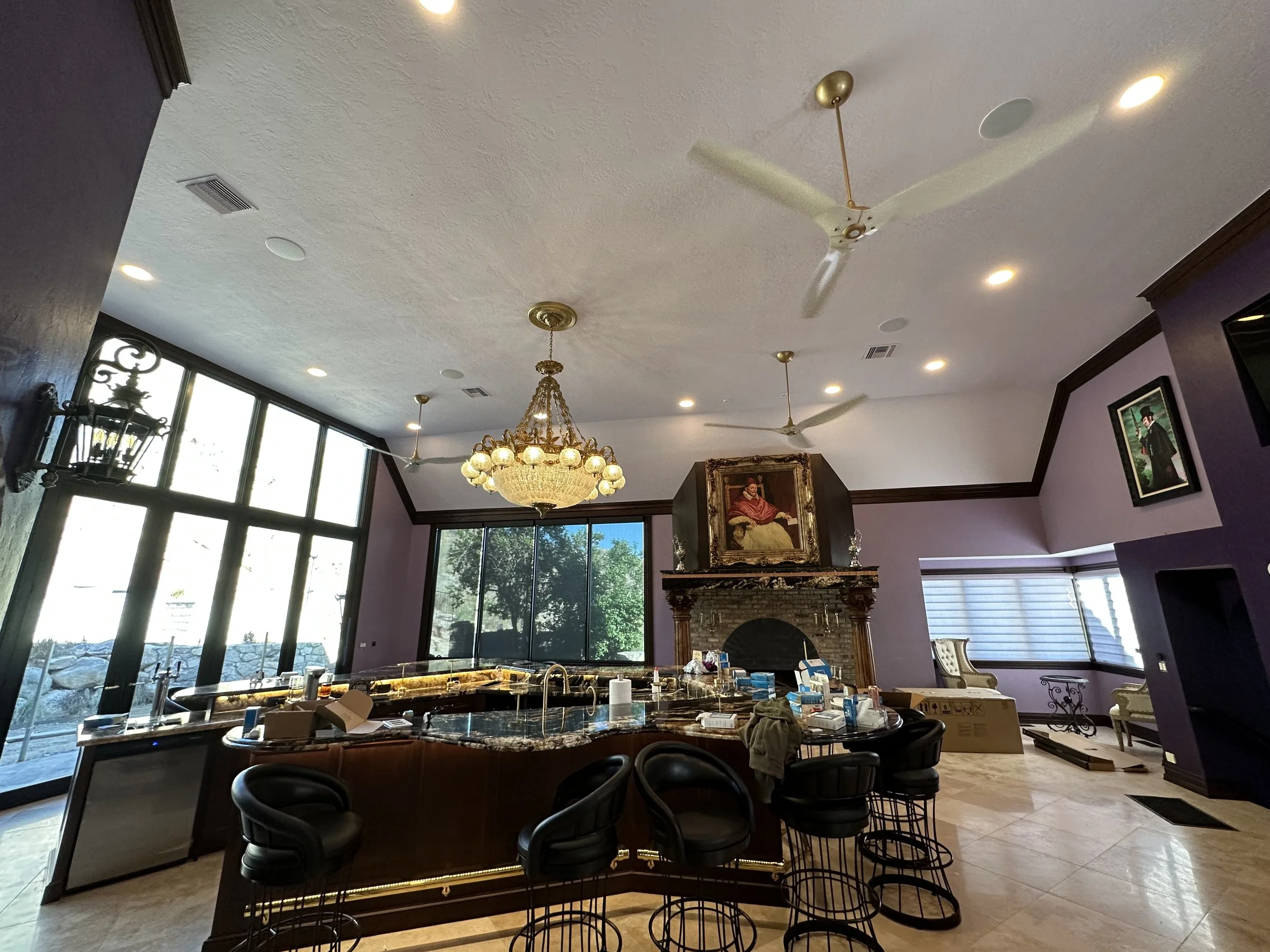 Interior of a spacious living room with large windows, purple walls, a chandelier, ceiling fans, a fireplace, and a curved kitchen island with barstools.