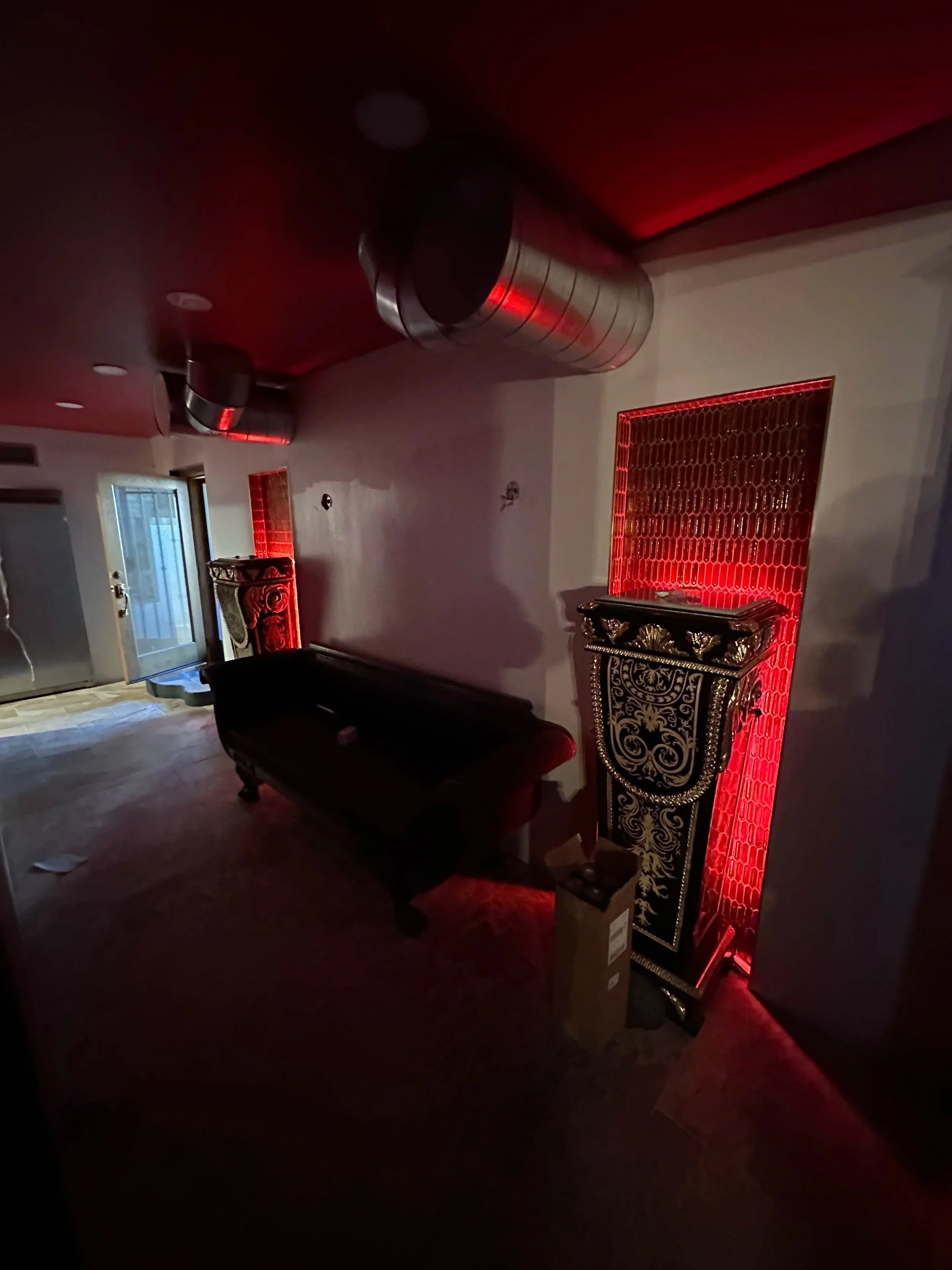 Dark room with red lighting, ornate black and gold furniture, large industrial silver air ducts on ceiling, and a partially open door leading outside.