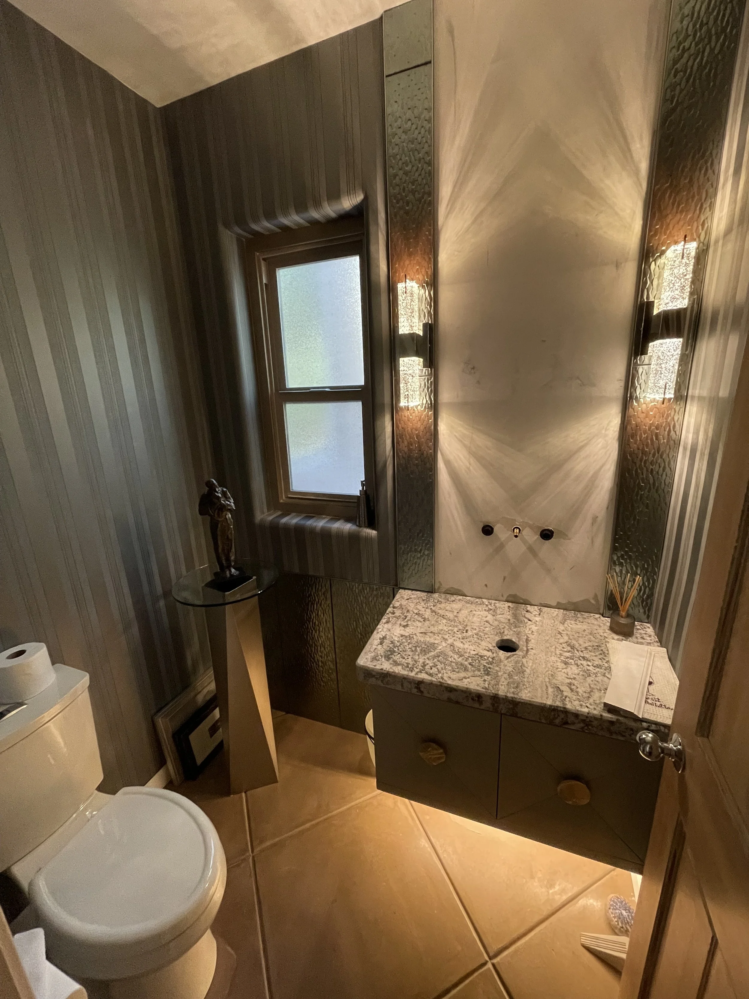 A small bathroom with a toilet, a marble-topped vanity, a mirrored wall with lighting, a window, and a small side table with a statue and flowers.