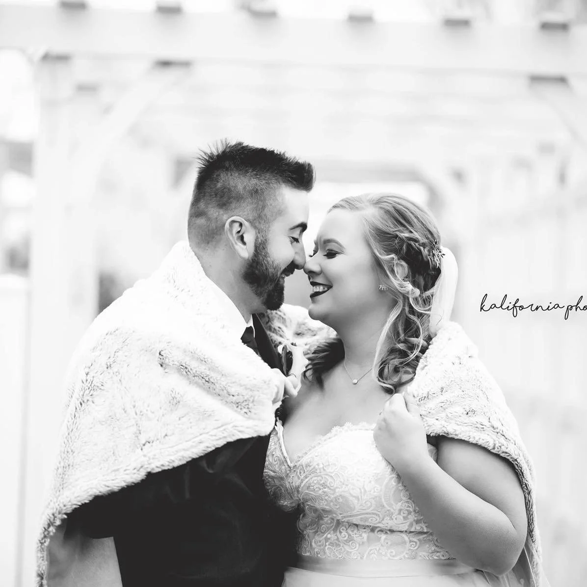 A black-and-white photo of a bride and groom smiling closely, with their foreheads touching, during a wedding celebration.