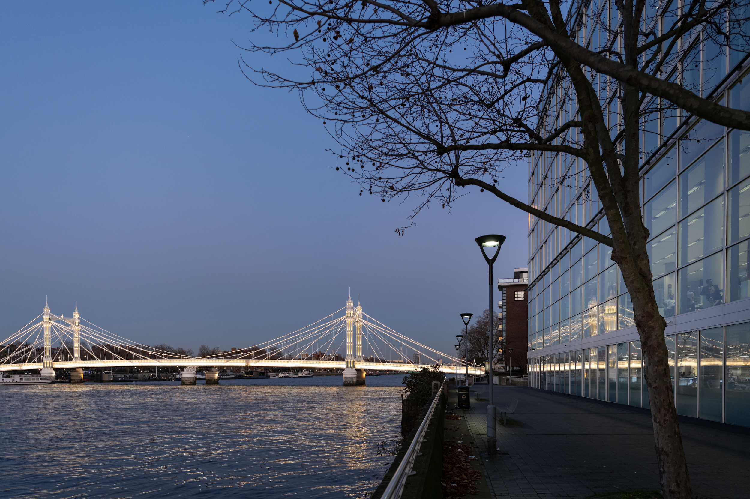 Photo of a cityscape at dusk showing a bridge illuminated with lights over a river, a modern glass building on the right, street lamps, and a leafless tree in the foreground.