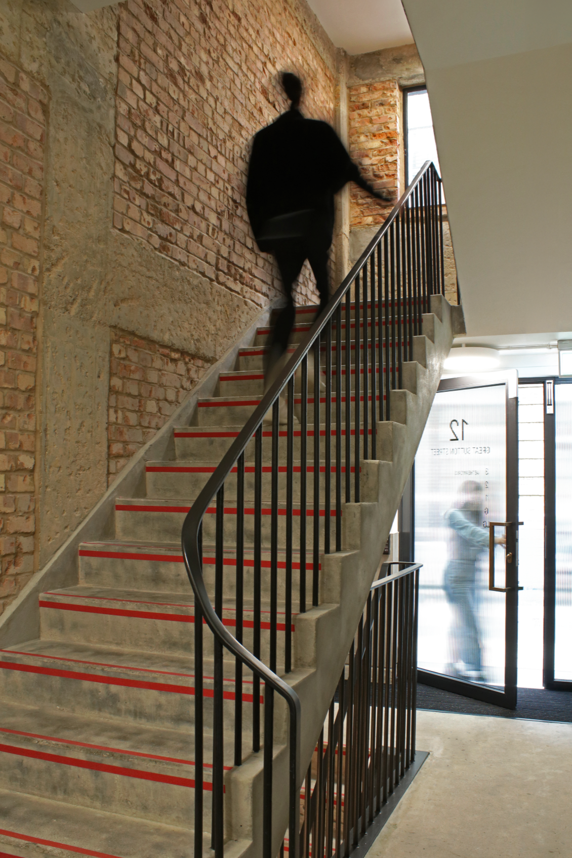 A person in black clothing walking up a zigzag concrete staircase with black metal railing inside a building with incomplete brick walls and a glass door.