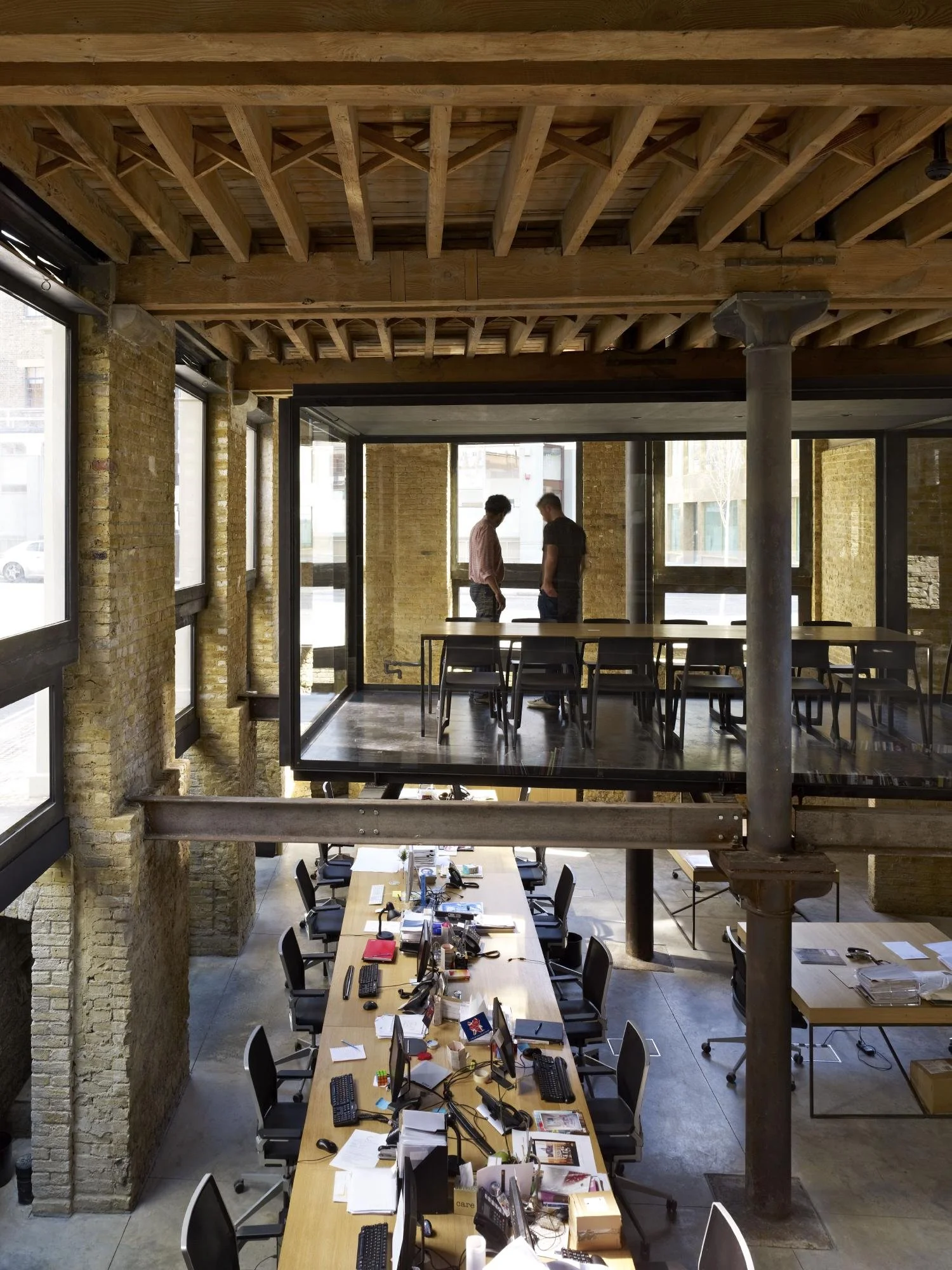 A spacious, industrial-style office with exposed brick walls, large windows, and a high ceiling with visible wooden beams. There are two levels connected by a glass balcony, with a group of two people talking on the upper level. The lower level has a