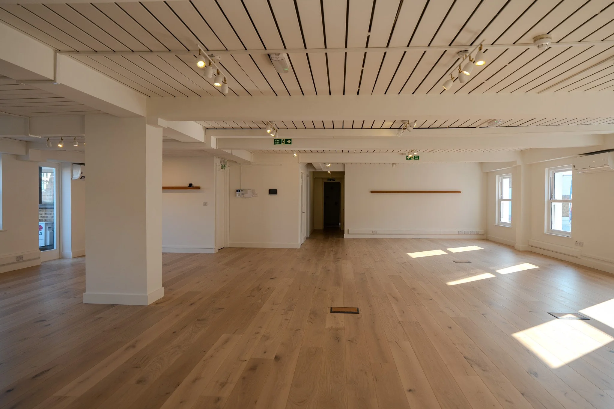 Empty, spacious room with wooden flooring, white walls, large windows letting in natural light, and ceiling track lighting.