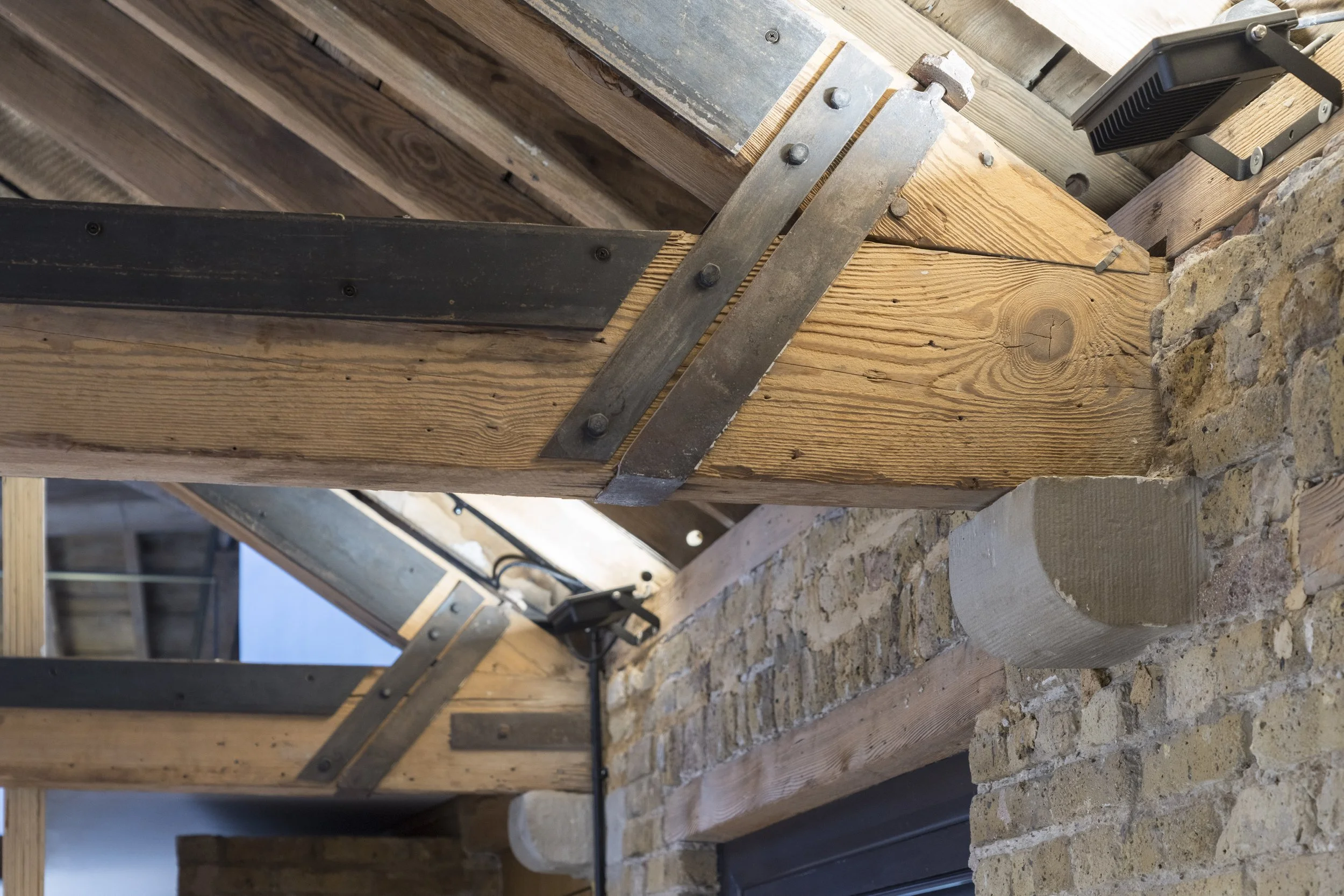 Close-up view of a construction site showing wooden beams, metal brackets, a brick wall, and a black electrical device mounted on the beam.