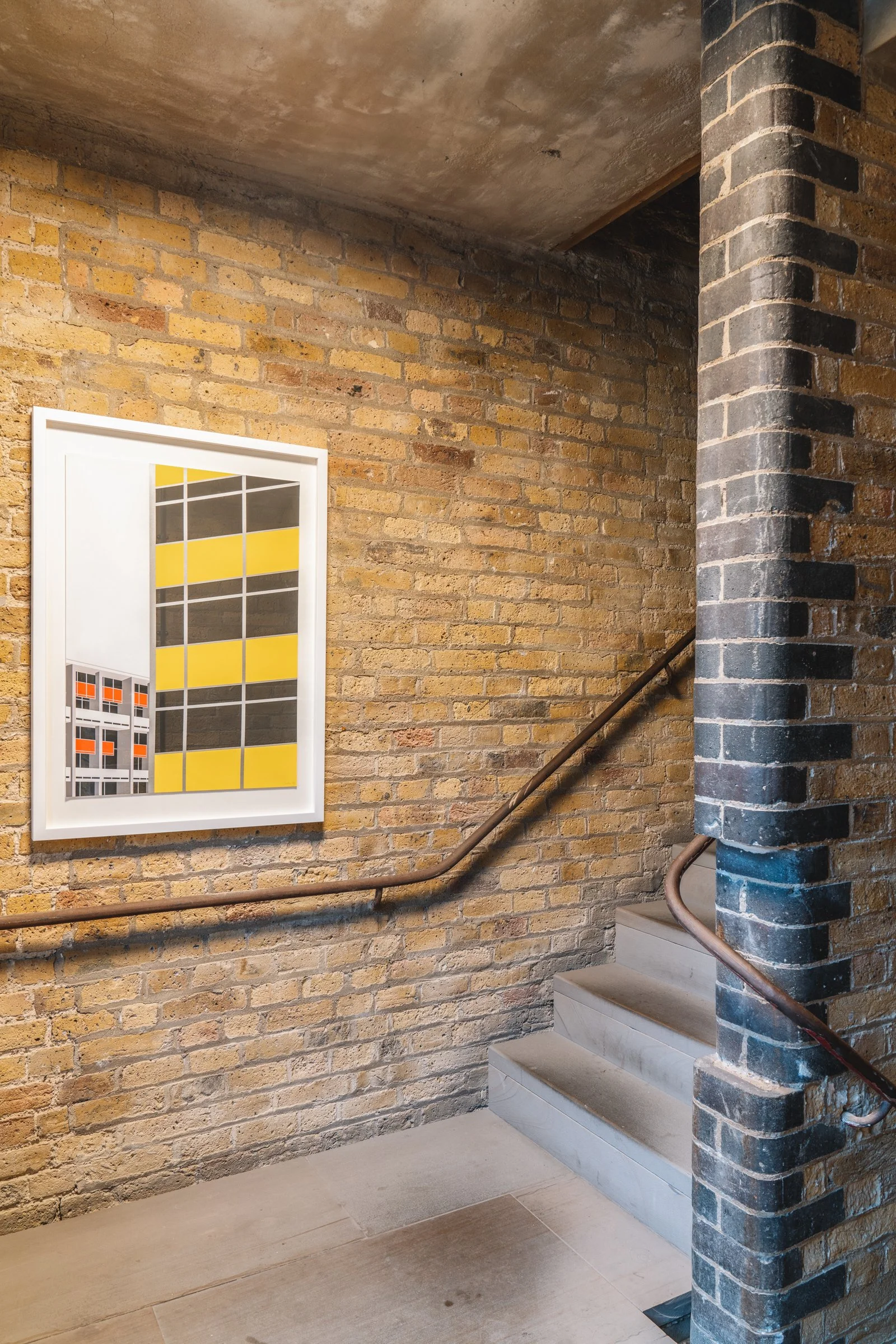 Inside a stairwell with exposed brick wall, a framed abstract painting with yellow, black, gray, and white geometric shapes, metal handrails, concrete stairs, and a brick column.