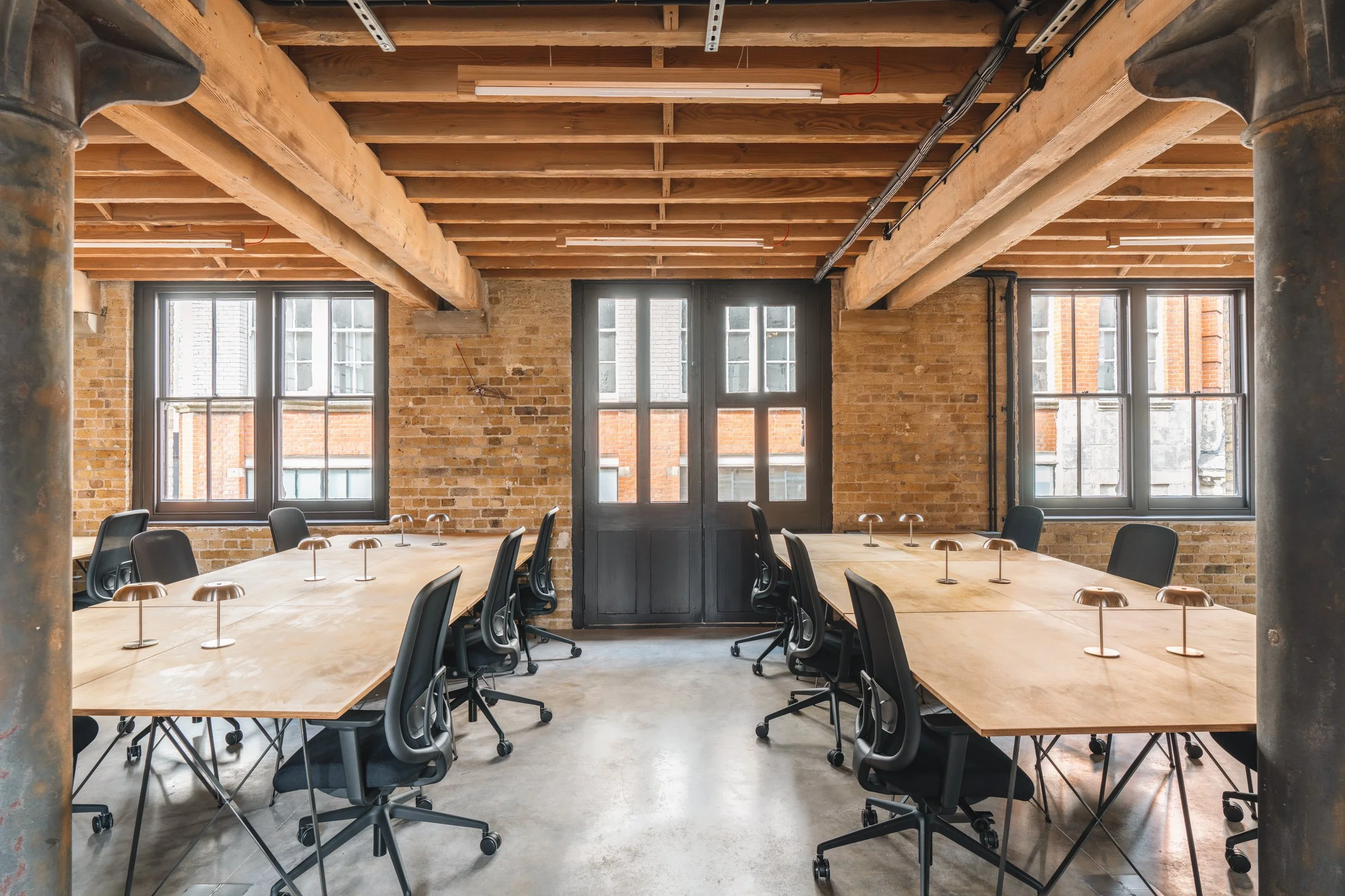 Empty industrial-style conference room with large wooden tables, black ergonomic chairs, exposed brick walls, and large windows.
