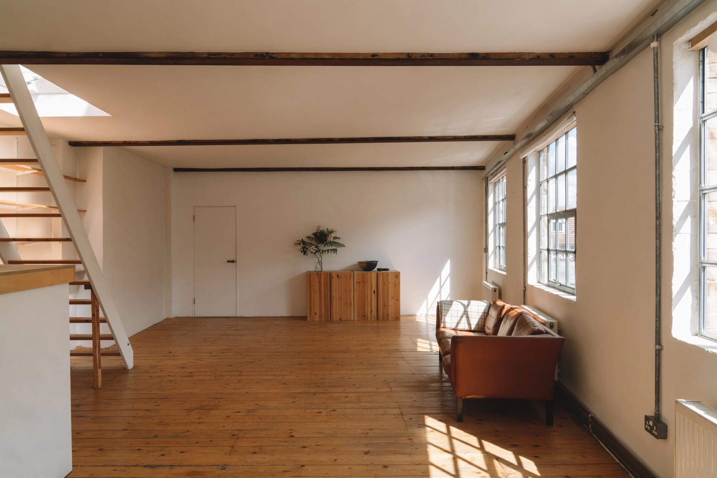 Spacious room with large windows, wooden floors, a brown sofa, and a wooden cabinet with a plant on top.