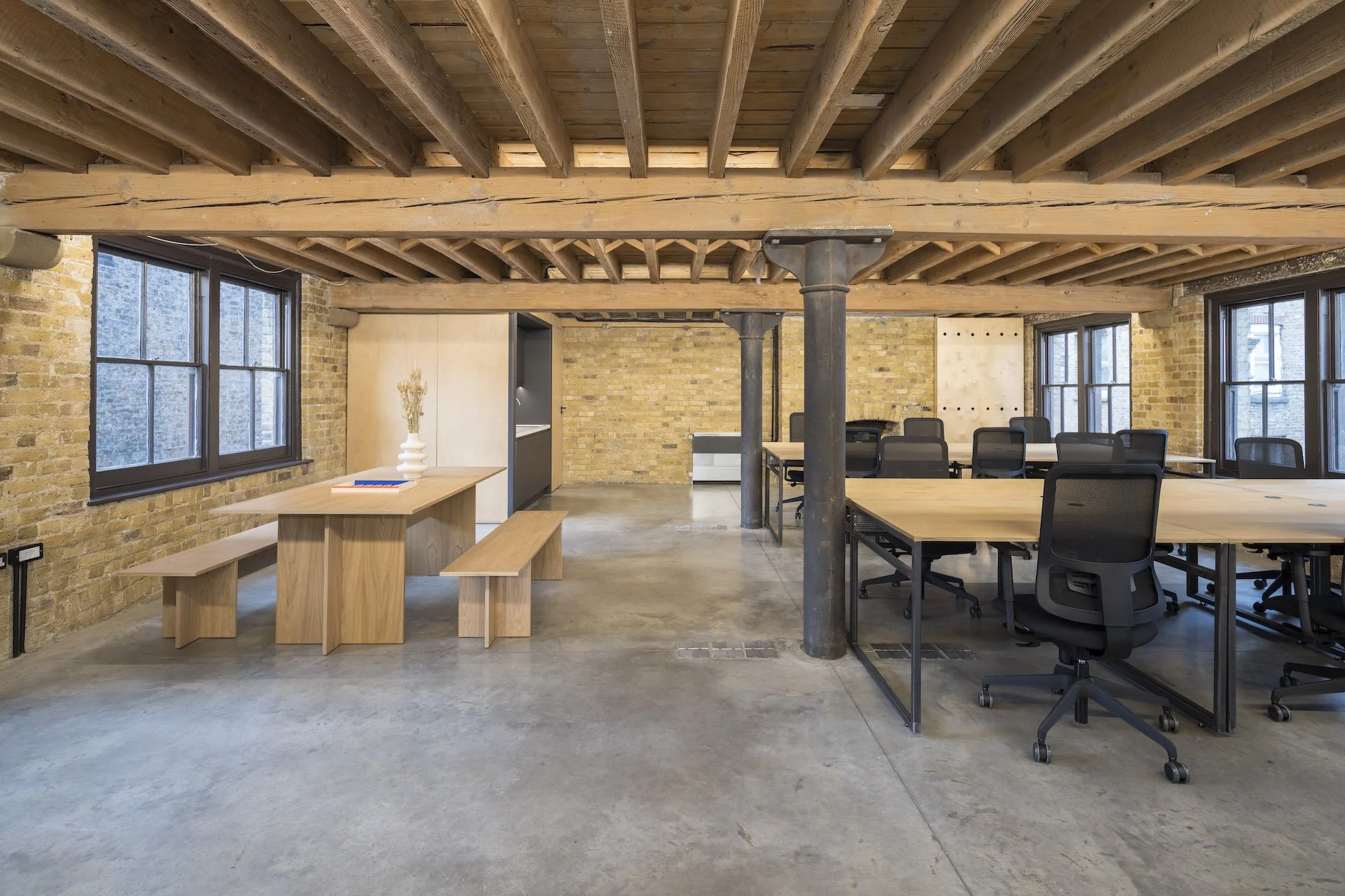 Modern office space with exposed brick walls, wooden beams, large windows, and contemporary furniture including a long table with chairs, a wooden bench, and a small table with a vase.