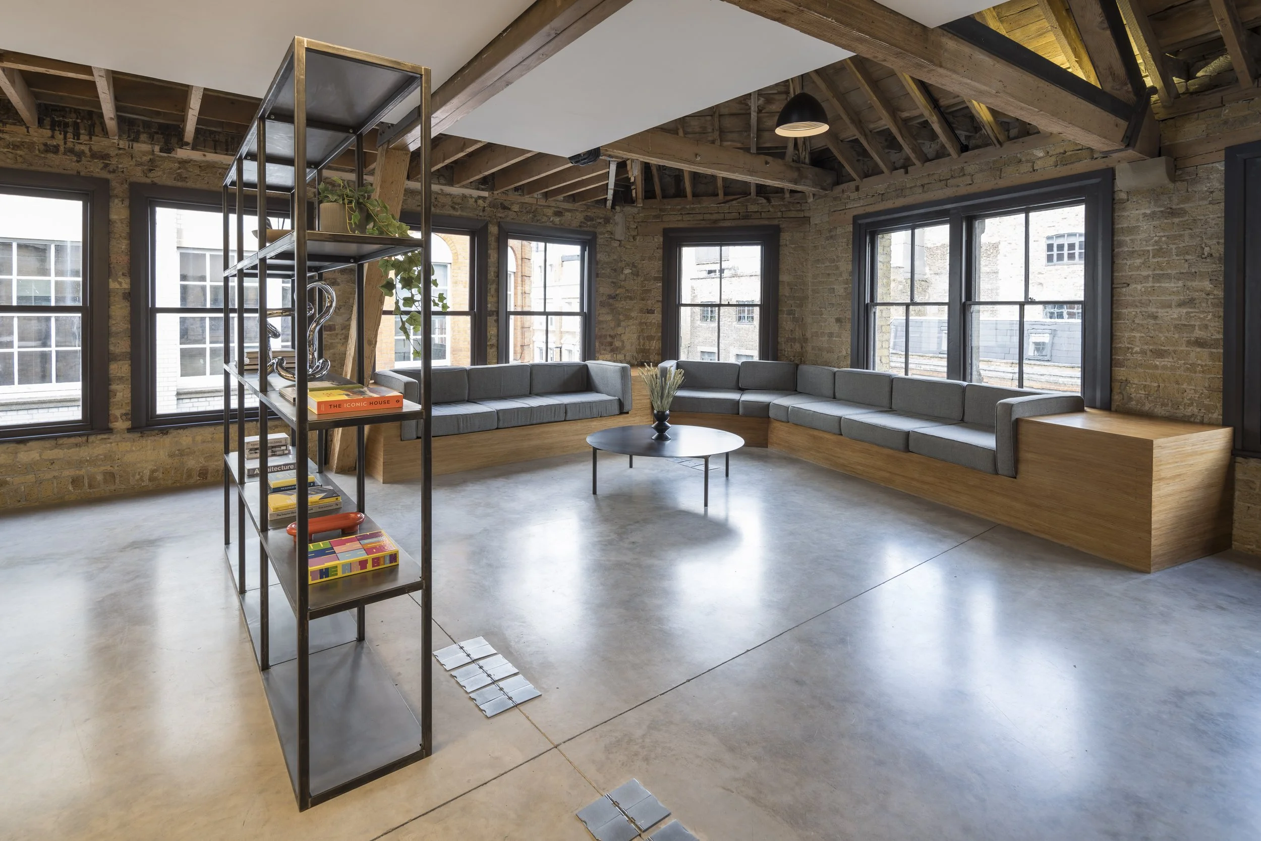 Loft apartment living room with exposed brick walls, large windows, a built-in gray cushioned bench with a wooden base, a black round coffee table with a vase of white flowers, and a black metal bookshelf with books and decor.