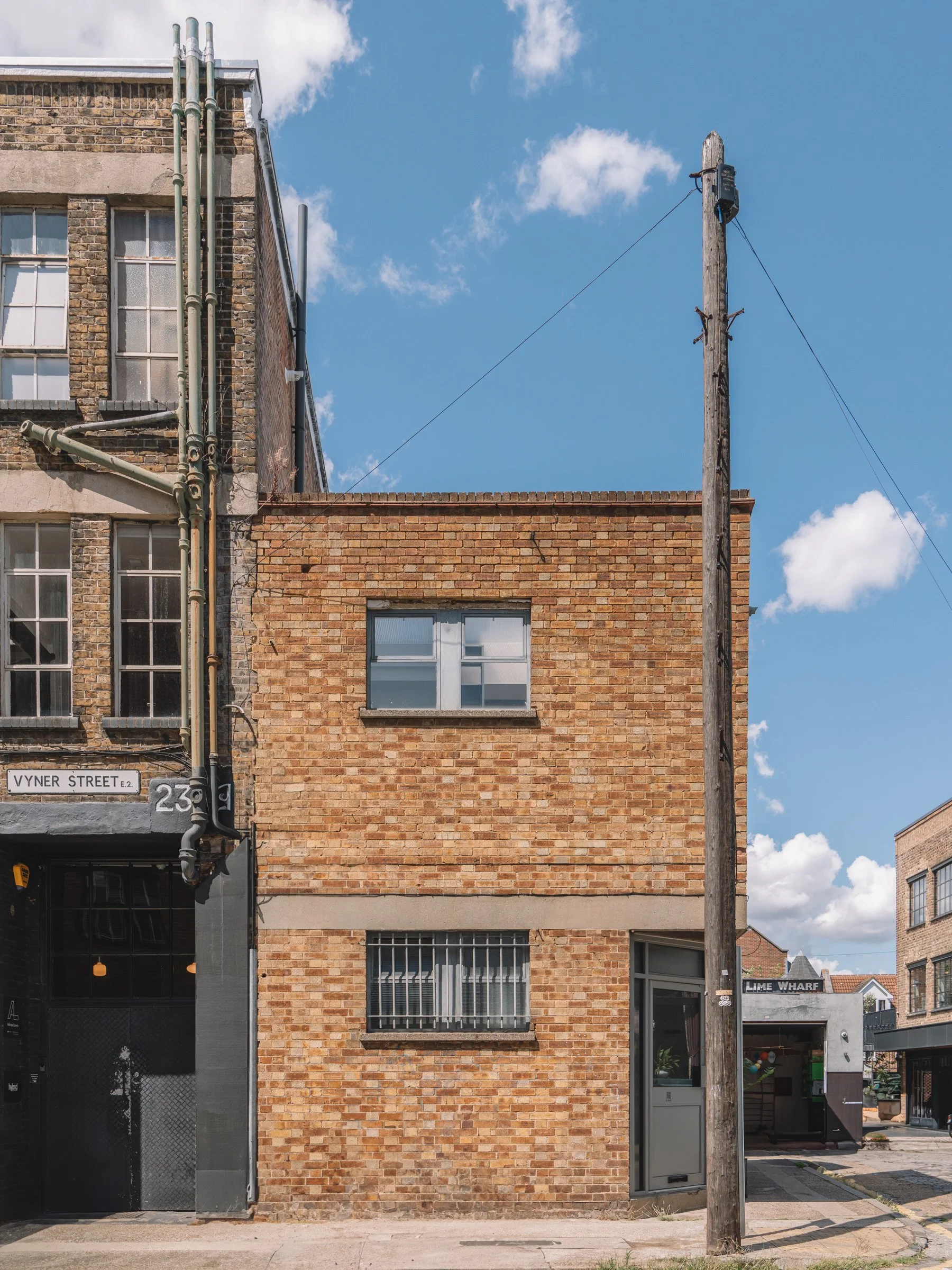 A small brick building with a metal door and barred window on the ground floor, and two small windows on the upper floor. There is a wooden utility pole with wires next to the building, against a backdrop of a partly cloudy blue sky.