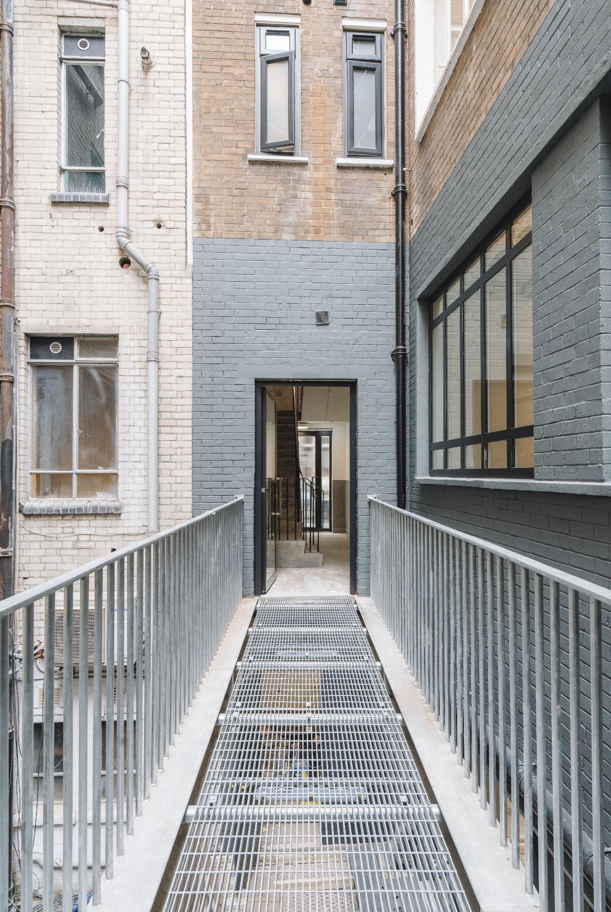 A metal ramp with railings on both sides leads up to a doorway in a building. The building has gray and brown brick walls, with large windows on the right and smaller windows on the left.