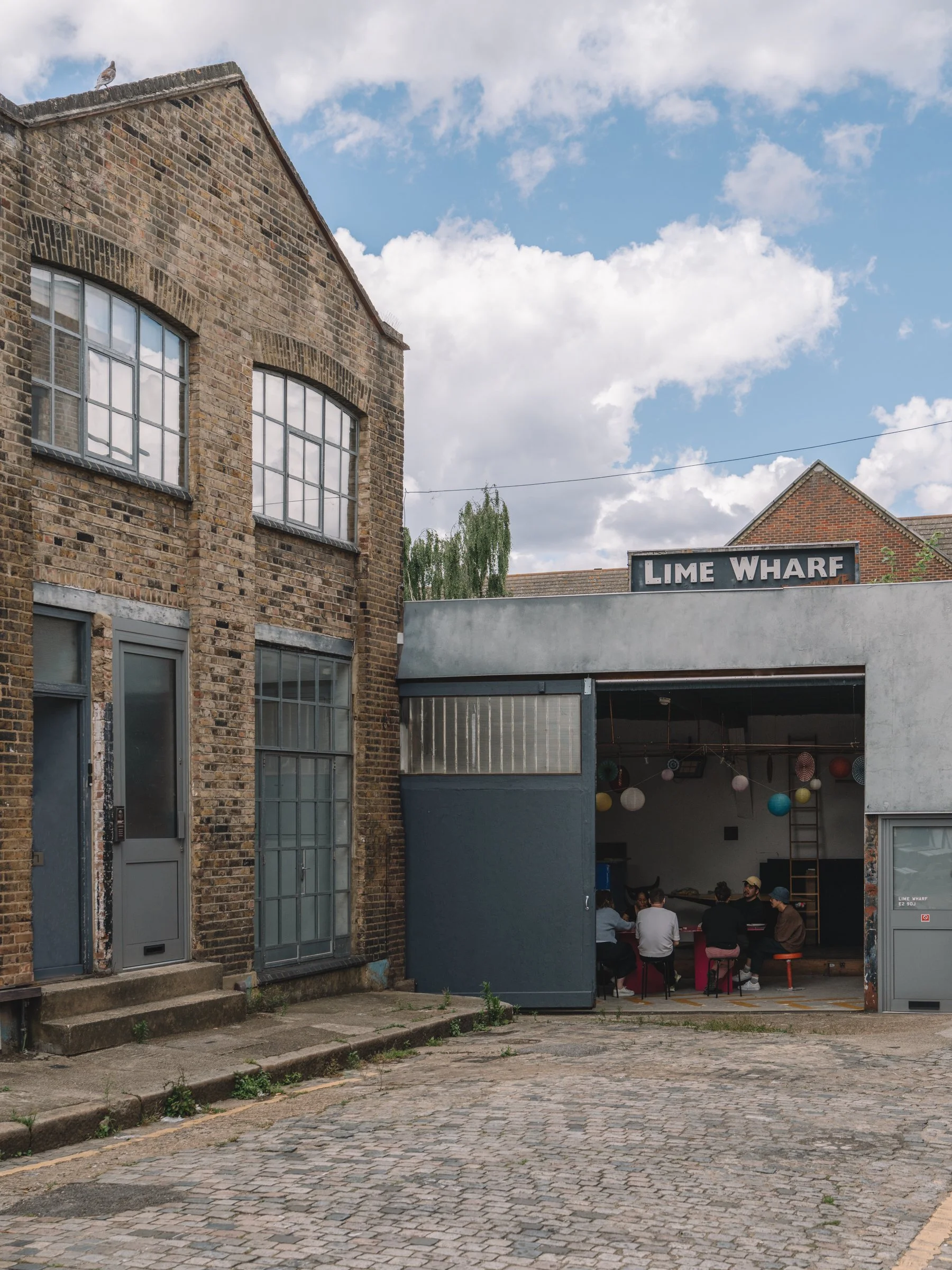 People sitting around a table in a garage-style venue with the sign 'LIME WHARF' above, decorated with colorful paper lanterns, with a brick building and cobblestone street outside.