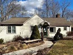 A single-story house with white siding, a brown shingled roof, and a small porch with steps. There are bushes and small trees in the front yard, and a stone walkway leading to the front door.
