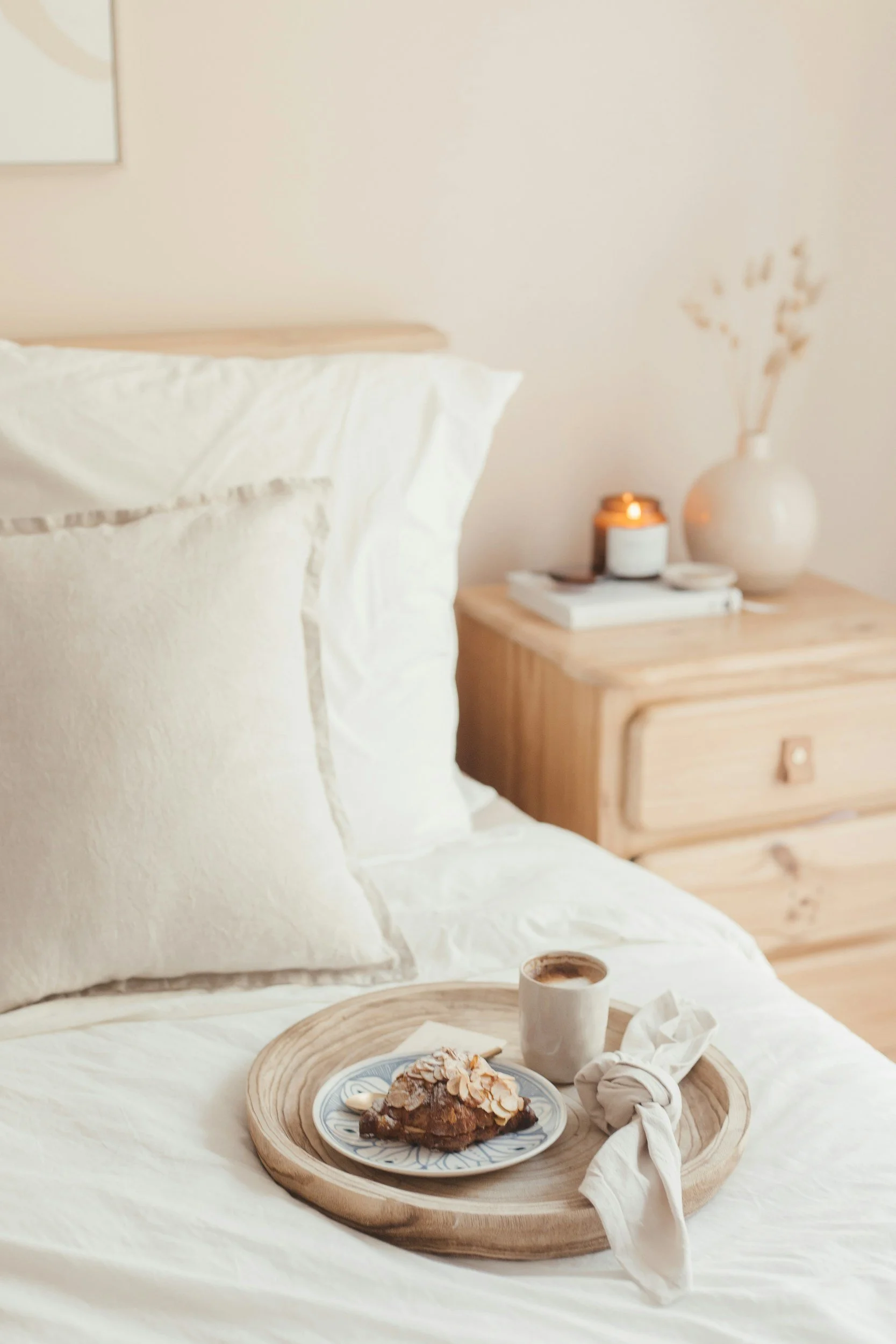 A cozy bedroom with white bedding and pillows, a wooden tray holding a pastry and a coffee mug placed on the bed, and a wooden nightstand with a lit candle, a vase with dried flowers, and books in the background.