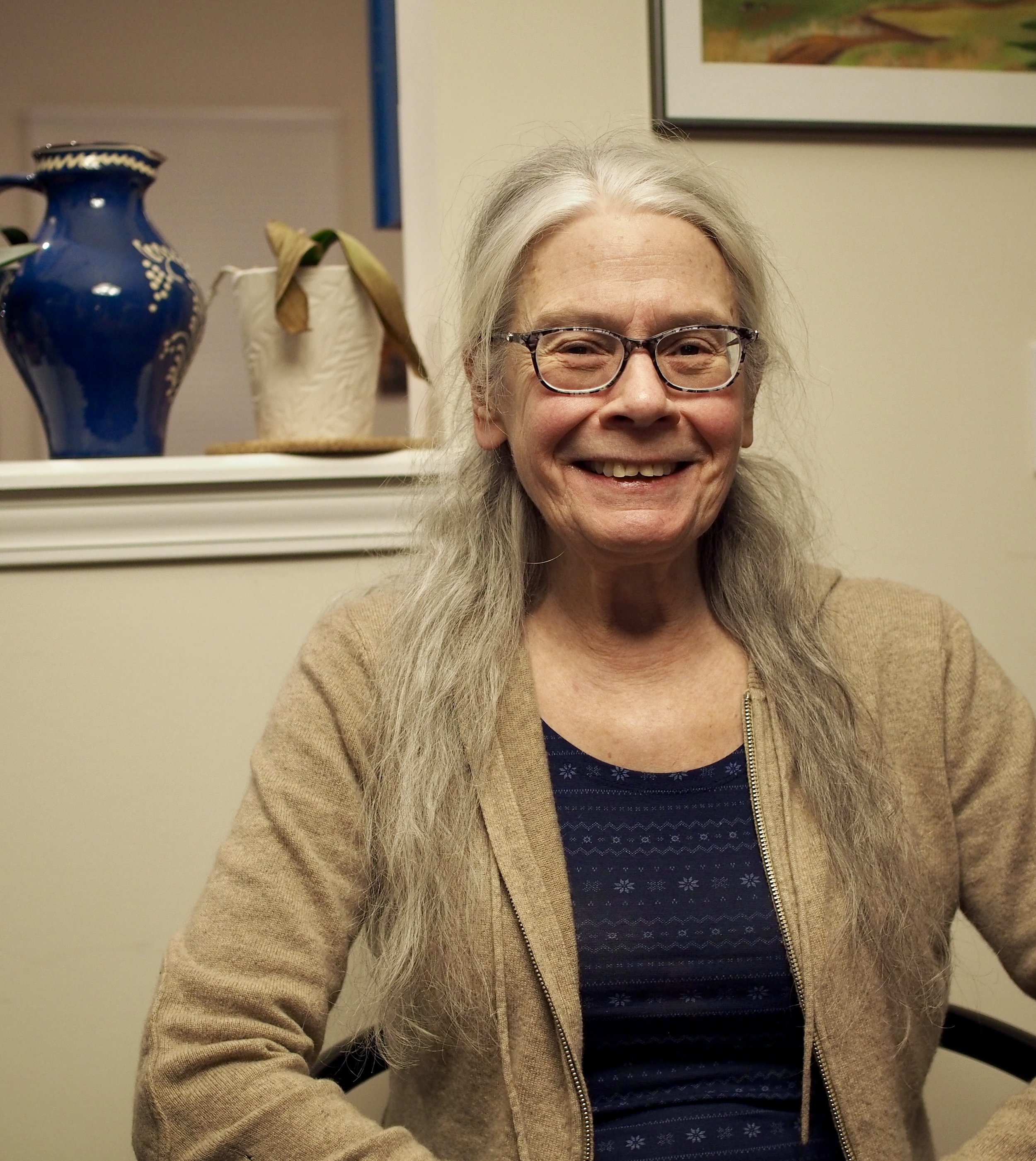 A smiling woman with long gray hair, wearing glasses, a beige jacket, and a navy blue shirt with a pattern, sitting indoors.