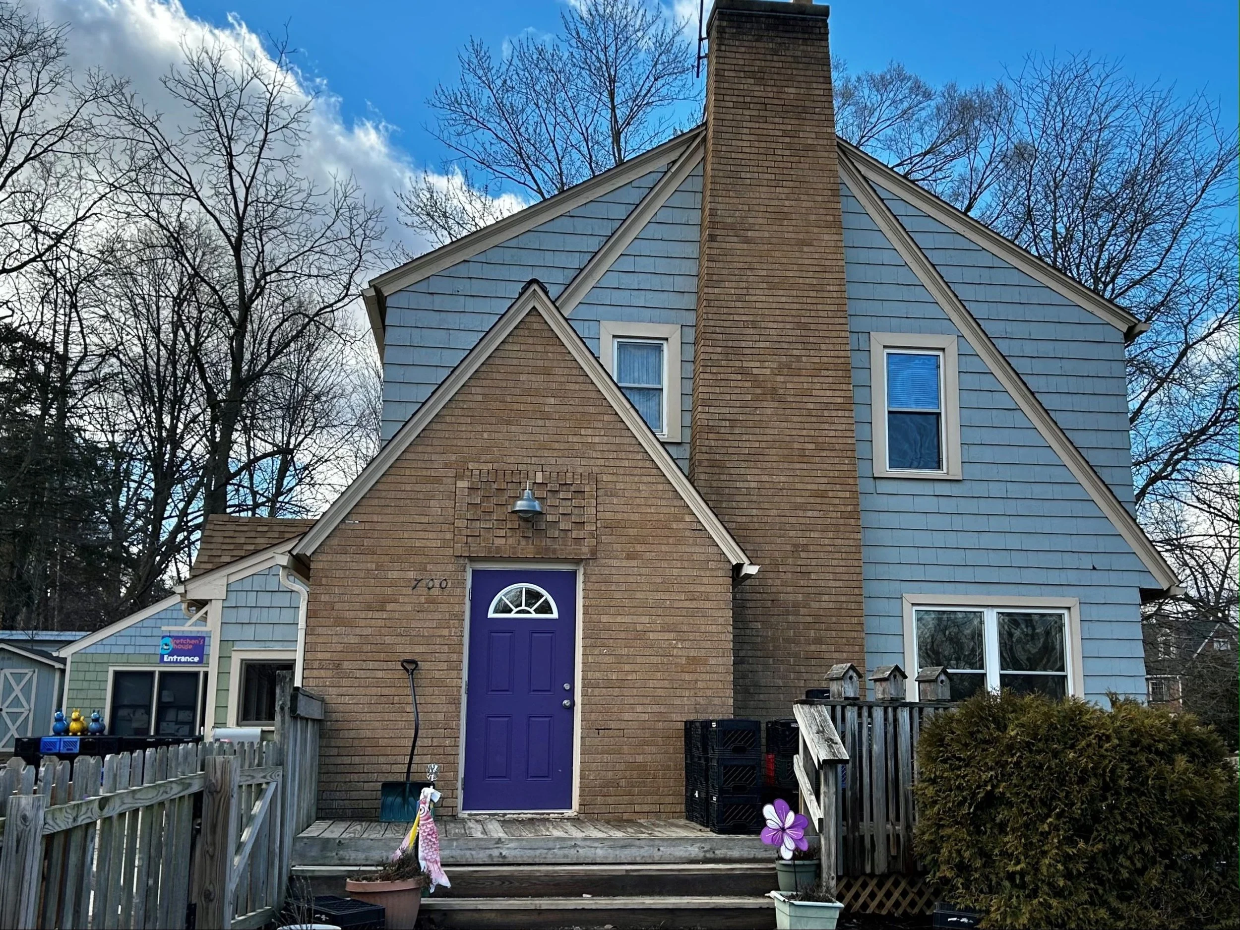 A two-story house with brick and blue siding exterior, a purple front door, and a chimney. The house has multiple windows, and is surrounded by leafless trees under a blue sky with some clouds.