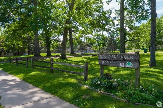 A park with tall trees, green grass, a wooden fence, a sidewalk, and a park sign that reads Elmendinger Park.