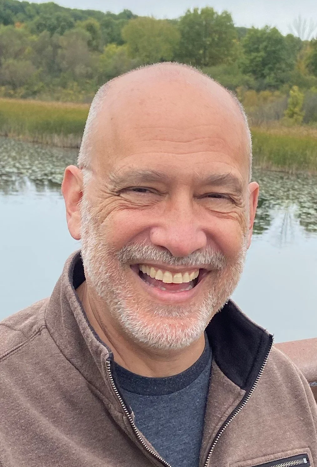 Close-up of a smiling man with a gray beard, wearing a brown jacket, outdoors near a lake with trees in the background.