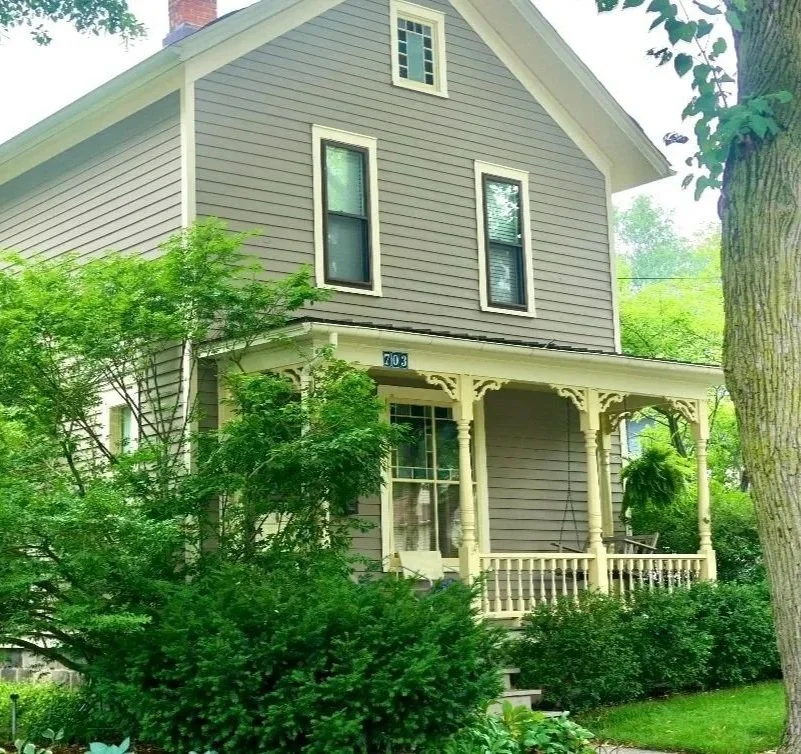 A two-story house with gray siding, white trim, and a front porch surrounded by greenery and trees.