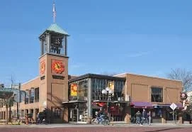 A city street scene with a building featuring a clock tower and a large emblem, with shops and pedestrians in front under a clear blue sky.