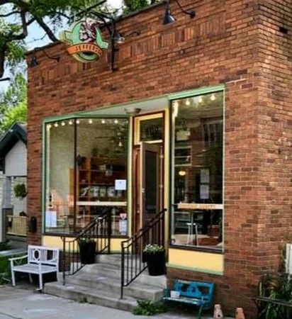 Front view of a brick storefront with large windows, a door, outdoor seating, and potted plants.
