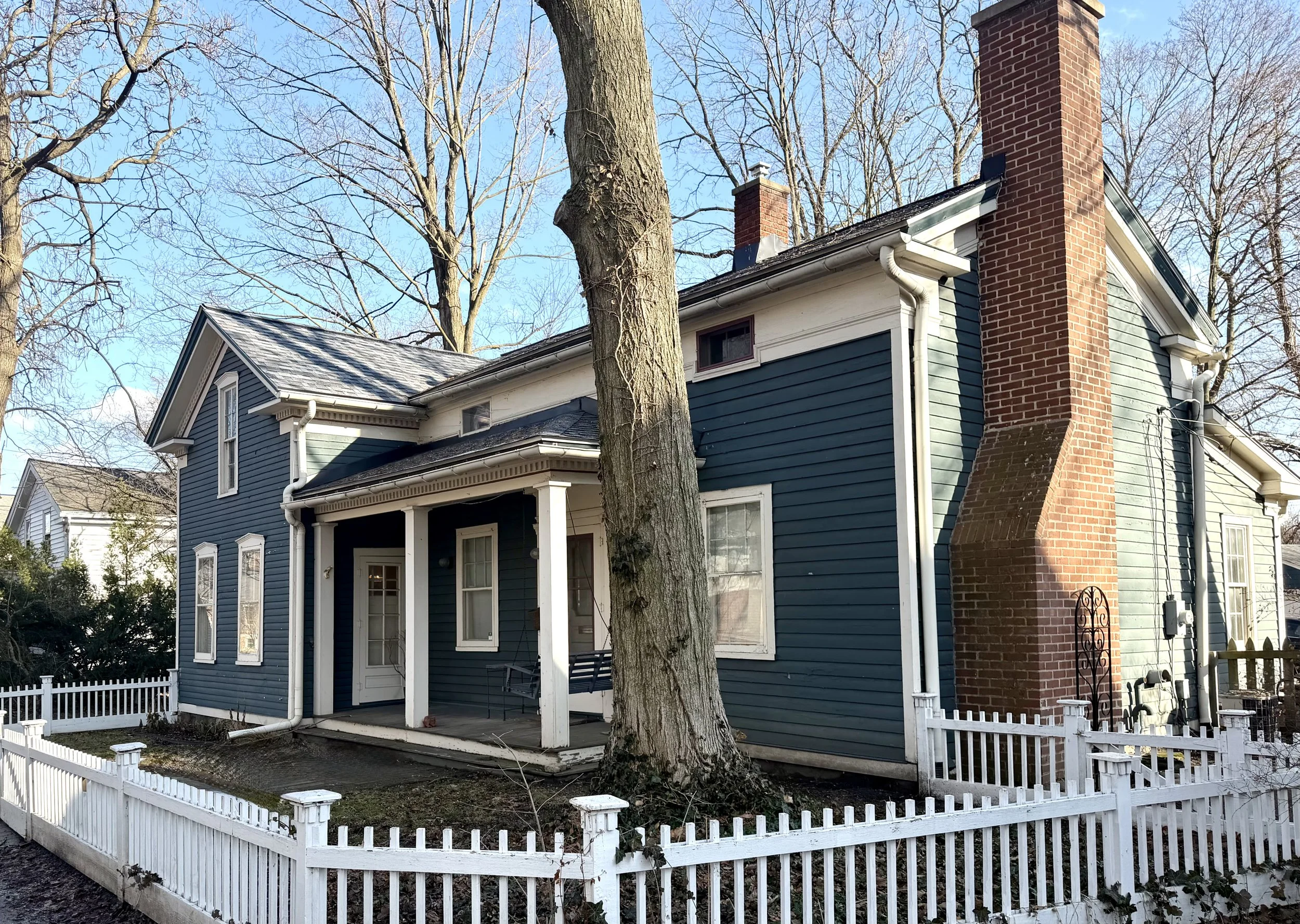 A blue house with white trim and a brick chimney, surrounded by a white picket fence, with a large tree in front and leafless trees in the background.