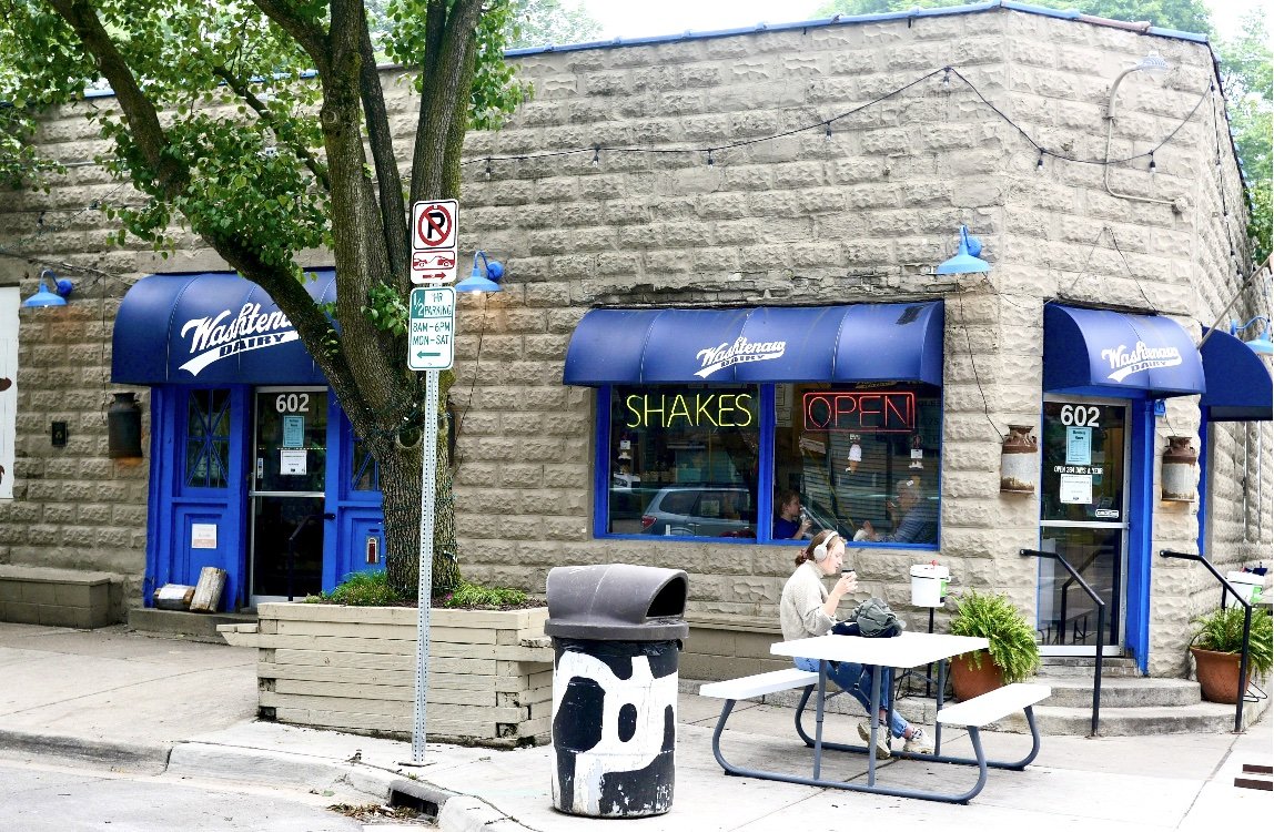 A small bakery named Wakenau Dairy with blue awnings and neon signs advertising shakes and that they are open. There is a person sitting at a picnic table outside, wearing headphones and looking at their phone. The building has a stone exterior and there are trees and street signs nearby.