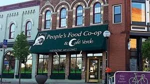 Exterior view of a building with a sign for People's Food Co-op & Cafe Verde, featuring trees and a bicycle rack outside.