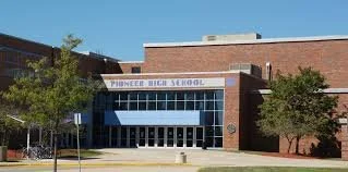 Exterior photo of Pioneers High School building with brick facade and glass entrance, trees in front, under a clear blue sky.