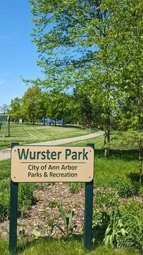 A park sign for Wurster Park in Ann Arbor, Michigan, with trees and a walking path visible in the background.