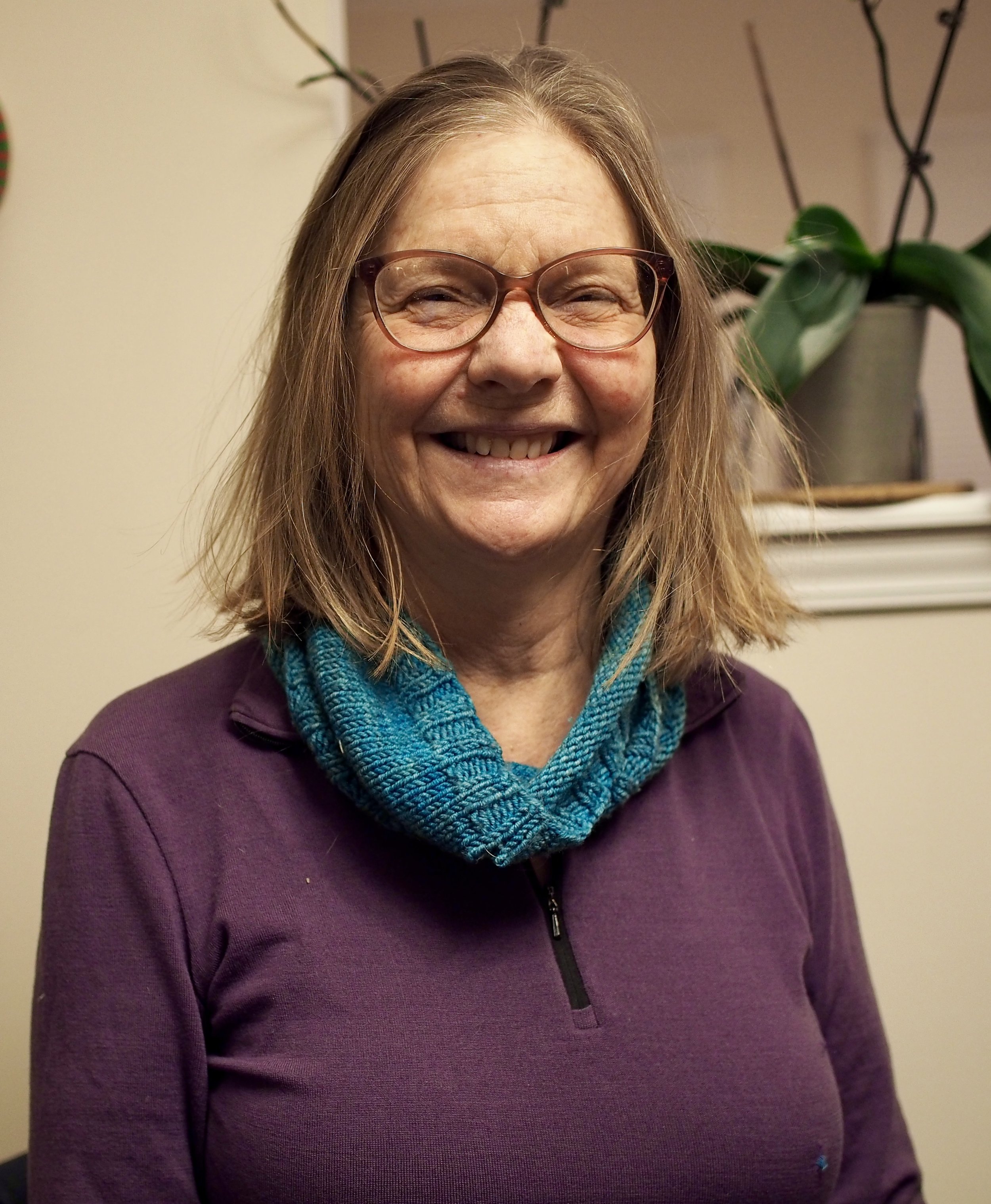 A cheerful woman smiling, wearing glasses and a purple top, with a bright blue scarf, standing indoors next to a houseplant.