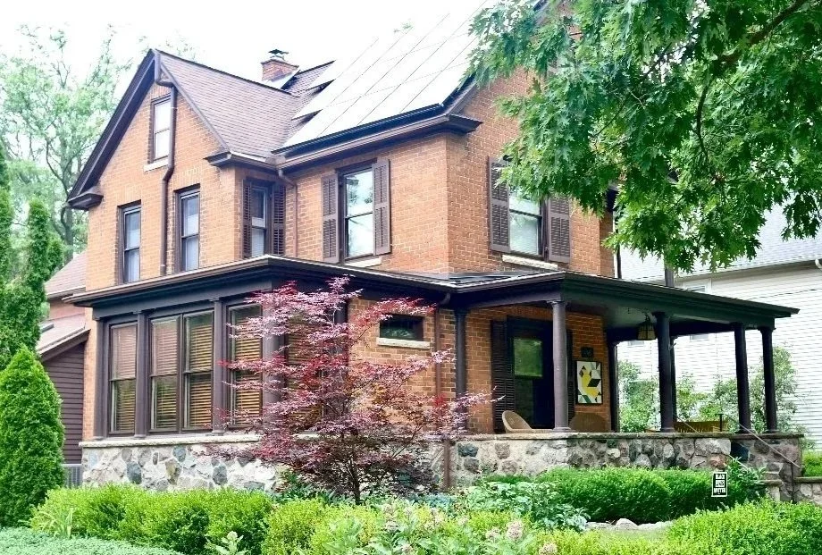 A two-story brick house with a covered front porch supported by black columns, surrounded by green trees and bushes, with a decorative quilt hanging on the porch wall.