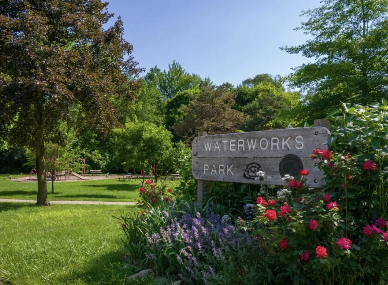 Park entrance sign for Waterworks Park with colorful flowers in the foreground and lush green trees and playground in the background on a sunny day.