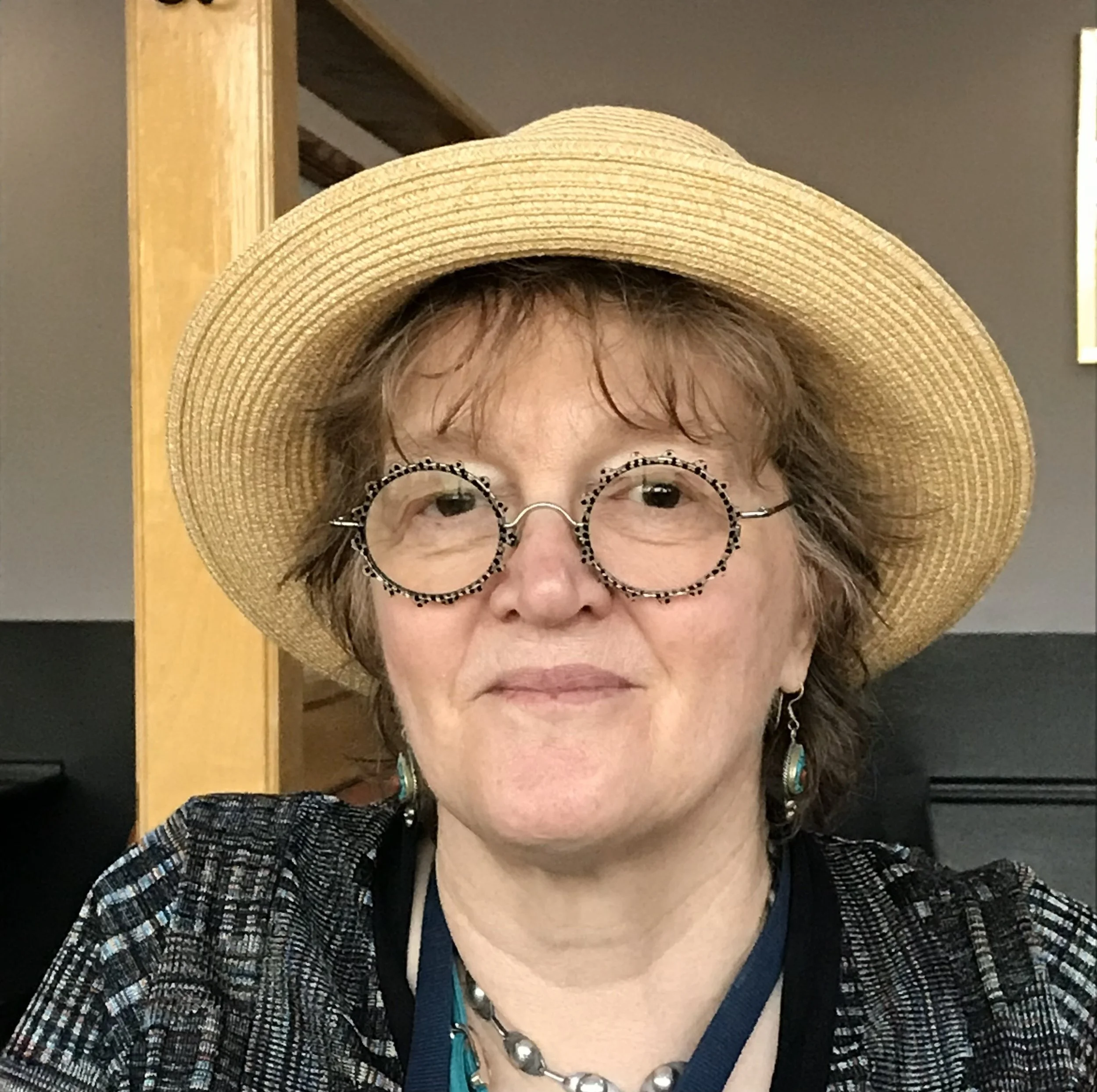 Woman with short brown hair wearing large straw hat, patterned glasses, earrings, and a necklace, smiling indoors.