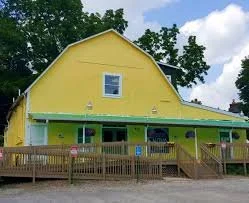 Yellow building with a porch and railing, set against a background of trees and a partly cloudy sky.