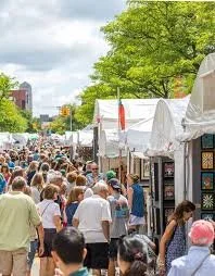 Outdoor street festival with white tents and a crowd of people, trees, and buildings in the background.