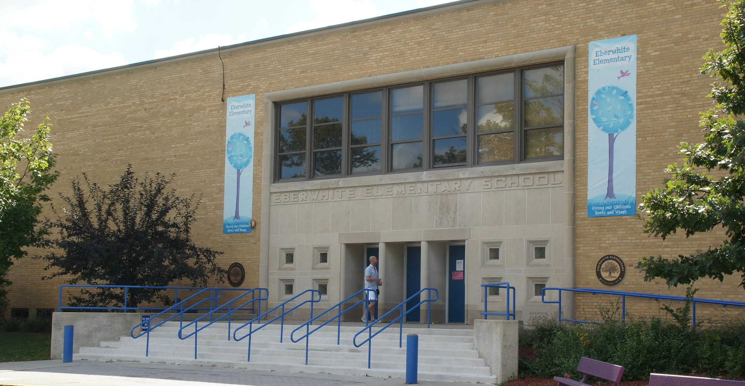 Front view of Eberwhite Elementary School with banners on the building, stairs with blue handrails, and a person standing near the entrance. Trees and a bench are visible in the foreground.