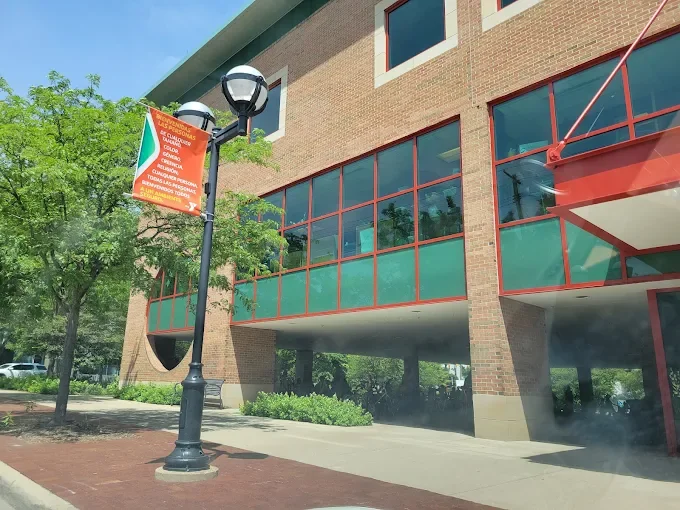 A brick building with large red-framed windows, a black lamppost with a colorful banner, and some greenery in front.
