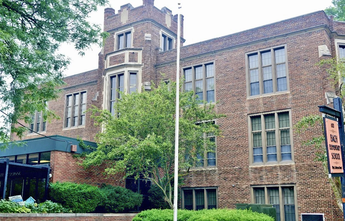Brick educational building with multiple windows, surrounded by trees and bushes, with a sign indicating it is a block elementary school.