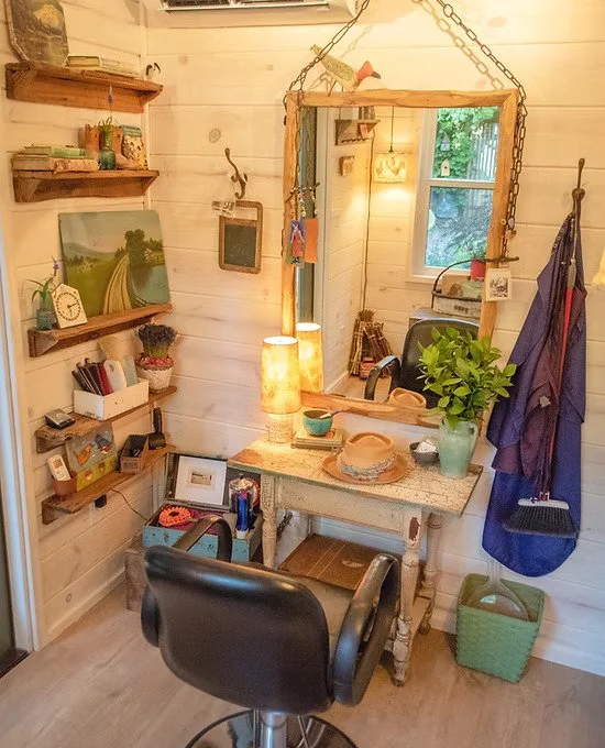 Cozy corner with a wooden vanity table, large mirror, chair, and decor including plants, lamps, shelves with books and art, and a window with a view of greenery.