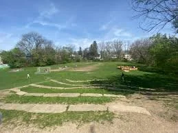 A park with paved pathways, grassy areas, and trees under a blue sky.